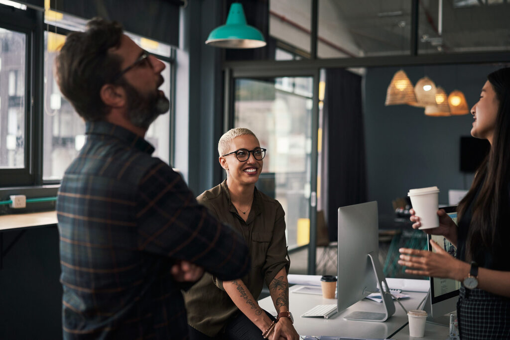 Three coworkers, two women and one man, in a break room with coffee, laughing with ease